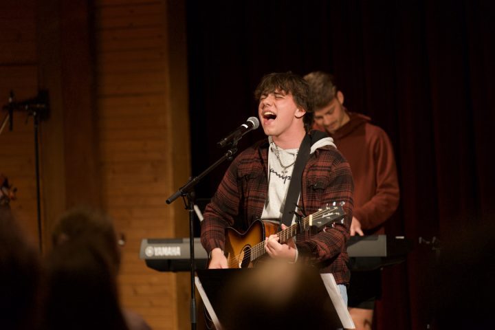 Picture of Malone student strumming a guitar and singing into a microphone on stage.
