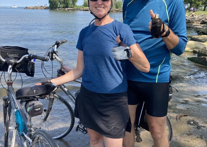 Ed and Brenda Walsh standing beside Lake Erie with their bicycles, both signaling a thumbs up