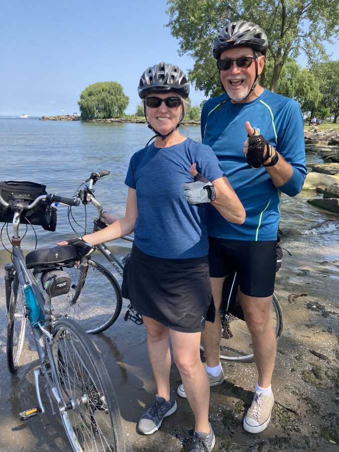Ed and Brenda Walsh standing beside Lake Erie with their bicycles, both signaling a thumbs up