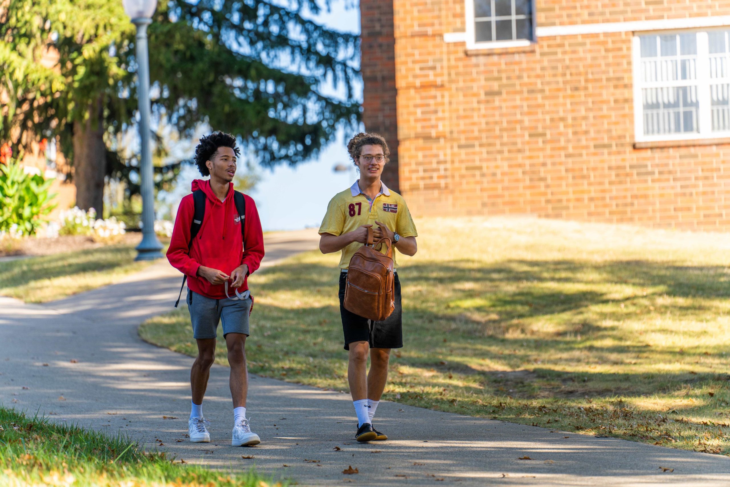 2 guys walking on a campus sidewalk; one carrying book bag, the other with his book bag strapped on