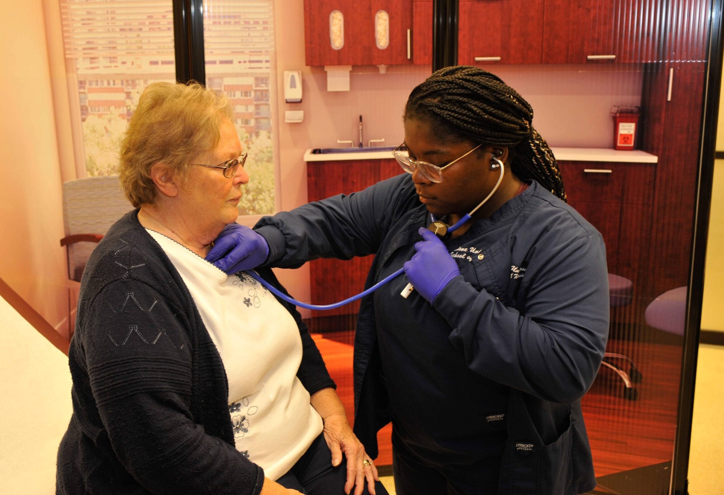 female nursing student dressed in blue scrubs listening to a patient's heartbeat
