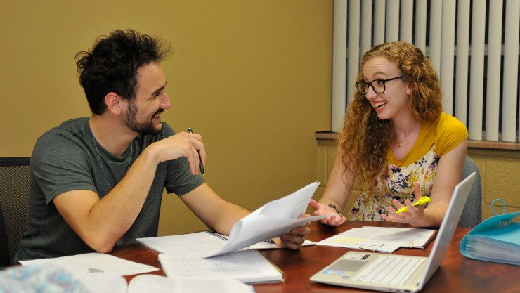 a guy and girl talking while sitting at a table with papers and laptop