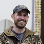 Daniel Bancroft smiling in a ball cap and camouflage jacket