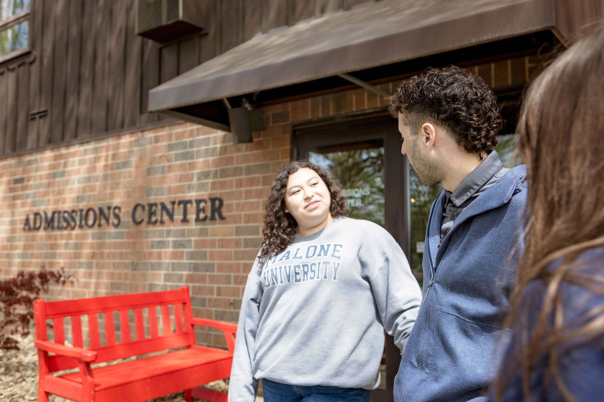 A female student with dark curly hair wears a Malone sweatshirt and talks with a male student outside the Admissions Center