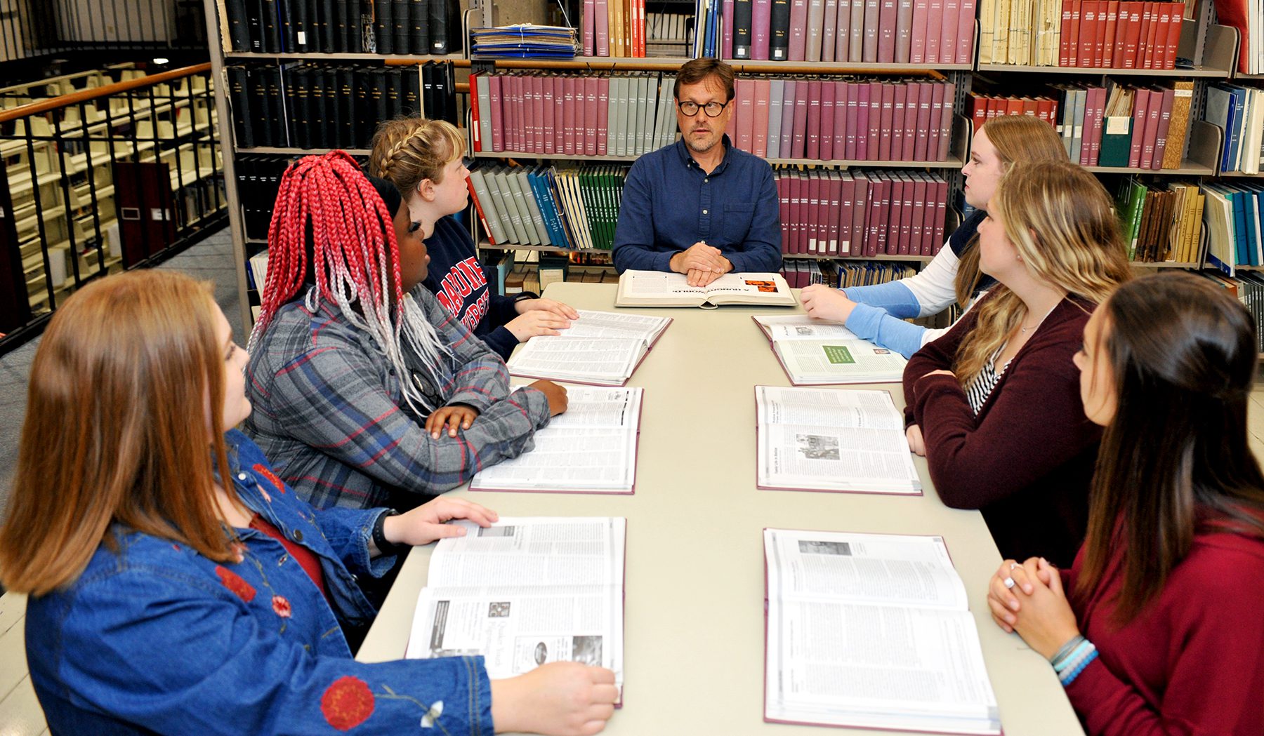 Students talking with professor sitting around a table with books in the background