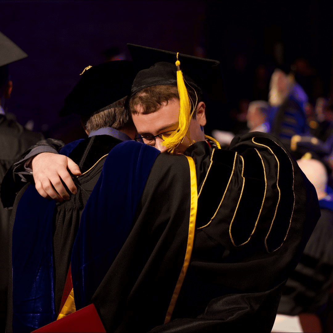 Male graduate in black cap and gown hugs a Malone administration member on stage at commencement