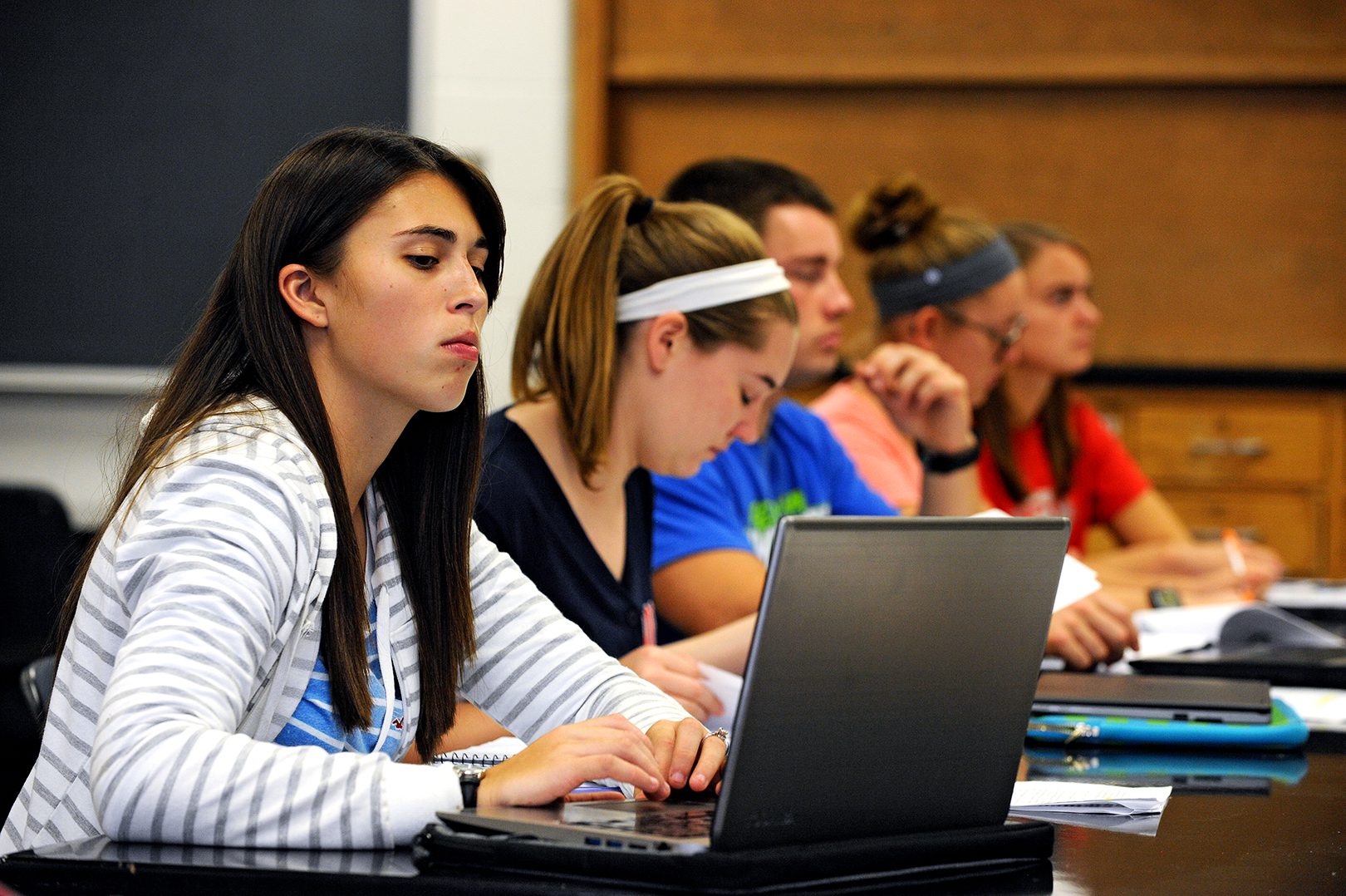 A female student takes notes on her laptop while sitting in a row of other students.